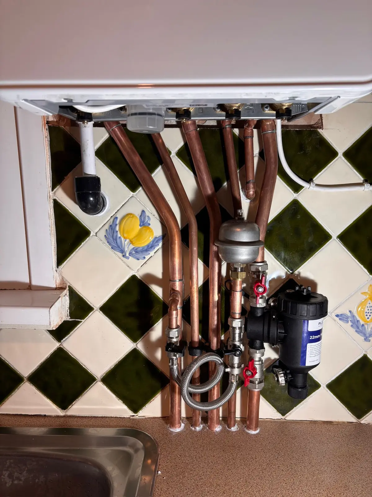 Copper pipework, isolation valves and a magnetic system filter neatly run beneath a combi boiler above a tiled kitchen worktop
